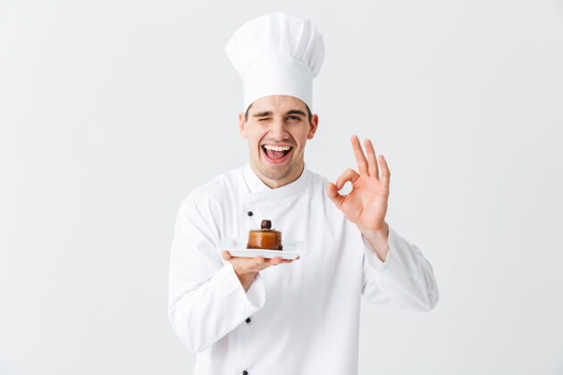 工人开朗的厨师穿着制服 在白色的墙上展示一个盘子上的糕点甜点男人厨房
