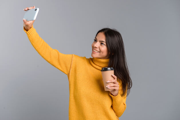 电话照片中 一位面带微笑的休闲年轻女子被隔离在灰色的墙上 手里拿着外卖咖啡 拿着手机自拍喝酒女孩白种人