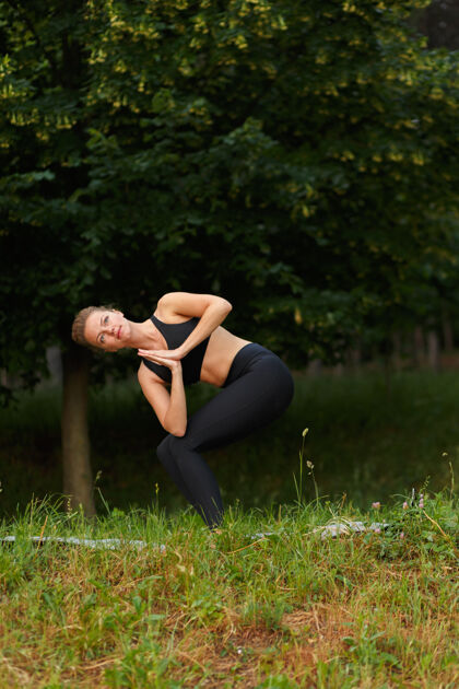 夏天一个穿着黑色运动服的年轻女子在公园里做瑜伽的全长肖像 森林公园背景.垂直查看锻炼女孩冥想