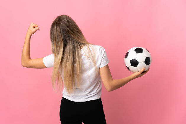 足球年轻的金发女郎拿着足球被隔离在粉红色的地板上休闲游戏女人