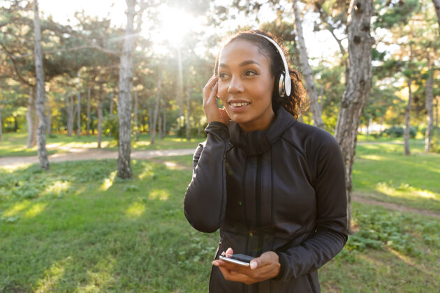 减肥20多岁的快乐女人穿着黑色运动服 戴着耳机 一边在绿色公园里散步一边用手机女性运动员公园