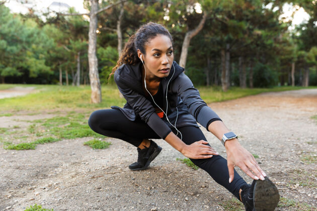 无线20多岁的美丽非洲裔美国妇女的肖像 穿着黑色运动服 在绿色公园里锻炼 伸展双腿吸引非洲健身