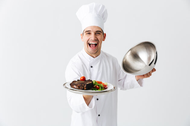 烹饪兴奋的男厨师穿着厨师制服打开便当盖隔离在白色的墙壁上 展示肉菜盖男性人