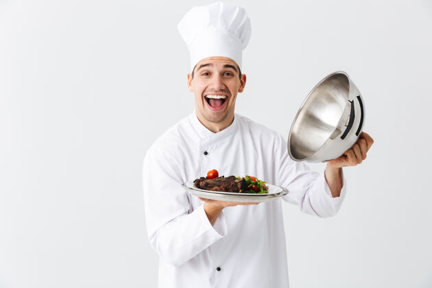 工作区兴奋的男厨师穿着厨师制服打开便当盖隔离在白色的墙壁上 展示肉菜帽子概念首席