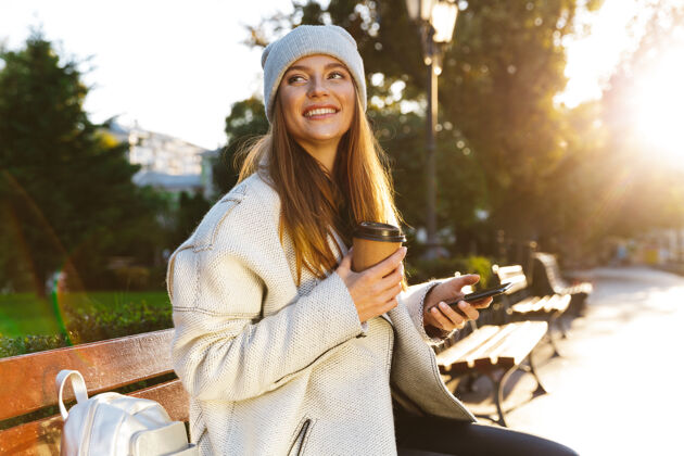 户外穿着秋衣戴秋帽的美女坐在户外的长凳上 喝着咖啡 用着手机人魅力早晨