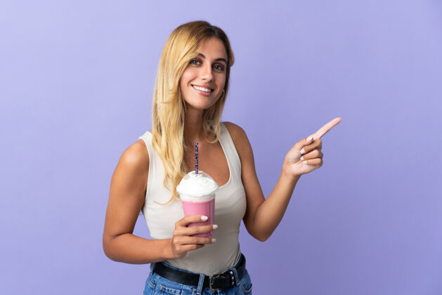 奶昔年轻的金发乌拉圭女人用草莓奶昔孤立地指着紫色的一面来呈现产品食物金发饮料
