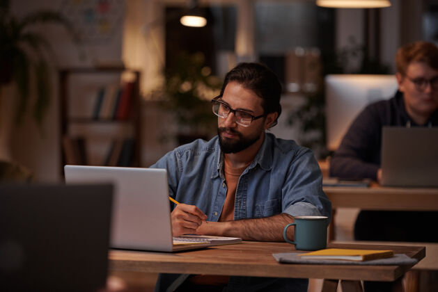 计算机年轻的留着胡子的商人戴着眼镜在办公室的桌子上做笔记和笔记本电脑坐着团队合作看样子