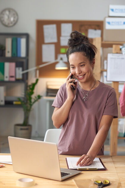 专业职业年轻微笑的女商人在手机上交谈 看着笔记本电脑 在办公室接订单裁缝通信计算机