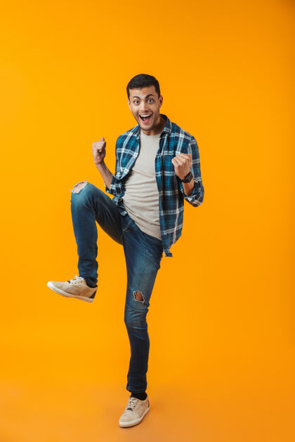 休闲一个穿着格子衬衫的快乐的年轻人的全长肖像被隔离在橙色的墙上 跳跃庆祝成功男站跳跃