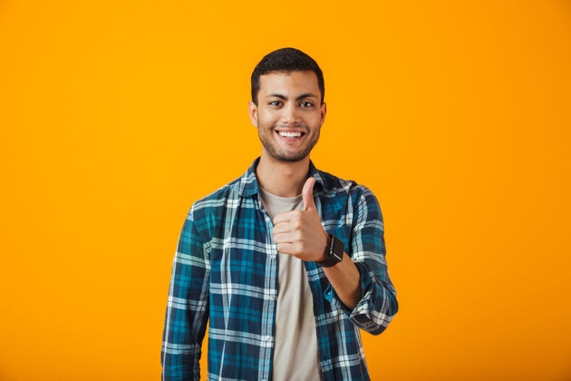 帅哥一个穿着格子衬衫的快乐的年轻人孤立地站在橙色的墙上 竖起大拇指男工作年轻