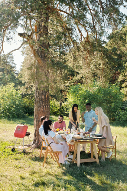 团聚三个年轻人站在餐桌旁 一边吃东西 一边喝饮料 而他们的朋友则坐在旁边 晚饭后聊天微笑野餐树