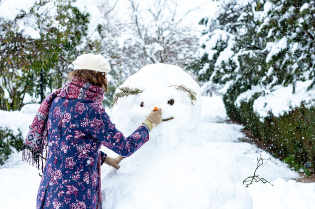 微笑一个穿着贝雷帽 夹克和围巾的金发女人在堆雪人的画像雪花冻结女性