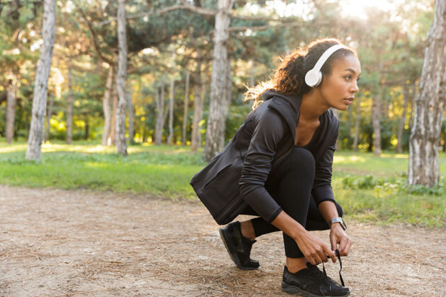 健身20多岁的黑发女运动员穿着黑色运动服系鞋带 在绿色公园里奔跑私人教练成人运动