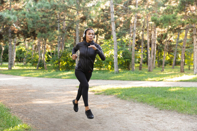 年轻一个20多岁的漂亮女人穿着黑色运动服 戴着耳机 一边在绿色公园里跑步跑步温暖伸展