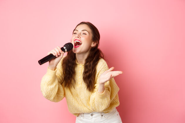 水平快乐的年轻女子喜欢唱卡拉OK 在麦克风里唱歌 一边看着屏幕上的歌词 面带微笑 站在粉红色的墙上黑发麦克风年轻