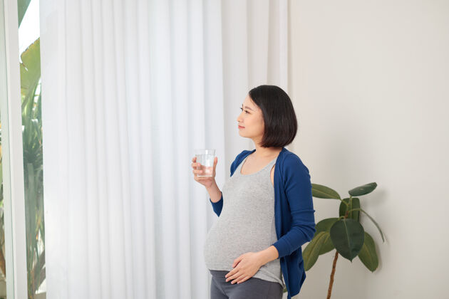 孕妇水化积极的孕妇饮用水从玻璃站在窗口看室内肚子女性肚子