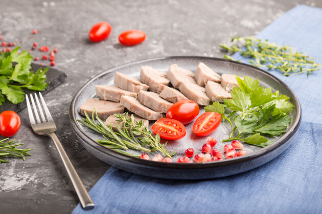 烹饪煮熟的猪肉舌头上有西红柿和草药的黑色混凝土表面和蓝色亚麻织物饮食肉小吃