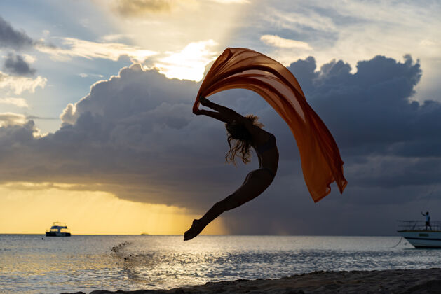 成功柔韧合身的女人在暴风雨云朵中戏剧性的日落中用丝绸跳跃机会自由海洋
