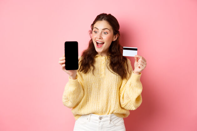 科技科技和网上购物兴奋迷人的女孩展示智能手机屏幕 塑料信用卡 看着手机 站在粉红色的墙上兴奋女性候选人