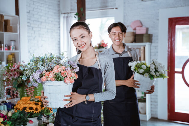 年轻快乐的年轻女花匠穿着围裙拿着桶花微笑着看着相机在花店工作的朋友在后面专业花卉室内
