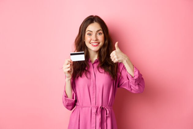 成人黑发女孩竖起大拇指 微笑着 推荐银行 拿着塑料信用卡 站在粉红色的墙上复印空间快乐候选人是