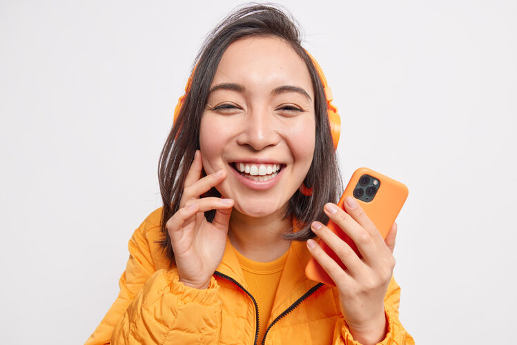 享受美丽开朗的亚洲女人的特写镜头从喜悦中绽放笑容欣赏喜爱的音乐手持身着橙色夹克的手机隔着白色的墙壁东方无线在线
