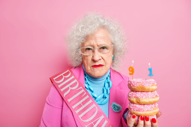 卷发摄影棚拍摄的满脸皱纹 头发卷曲的女人表情严肃 手里拿着一堆美味的油炸圈饼 穿着节日的衣服堆蛋糕粉色