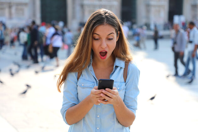 惊喜惊讶的年轻女子在户外使用智能手机城市街道电话
