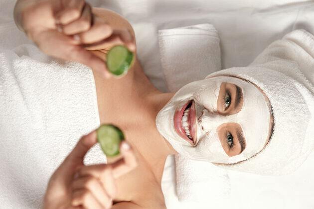 女孩美丽的温泉脸上戴着泥面具 手里拿着黄瓜的女人脱皮面膜.皮肤小心专业皮肤护理治疗