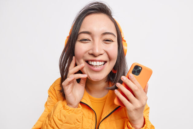 享受美丽开朗的亚洲女人的特写镜头从喜悦中绽放笑容欣赏喜爱的音乐手持身着橙色夹克的手机隔着白色的墙壁东方无线在线