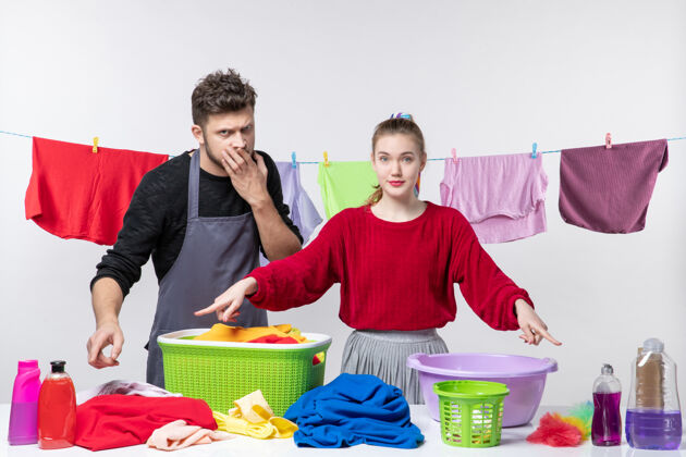 男人管家丈夫和妻子的正视图 他们指着白墙上桌子上的洗涤剂洗衣篮和清洁用品房子成人男性