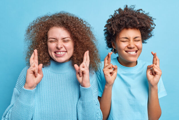 脸多民族妇女站在一起互相交叉手指祝好运预期好消息或结果闭上眼睛摆出乐观的姿势随意隔离在蓝色的墙上快乐人多样性