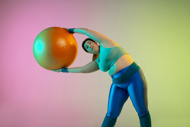 混合年轻白种人大码女模特在霓虹灯下的渐变紫绿色墙壁上训练健身房人积极