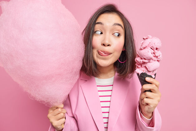 蛋卷美丽的亚洲女人舔着嘴唇看着开胃的冰淇淋拿着甜甜的棉花糖想吃美味的甜点穿着正式的衣服隔着粉色的墙壁是时候吃点美味了人美味室内