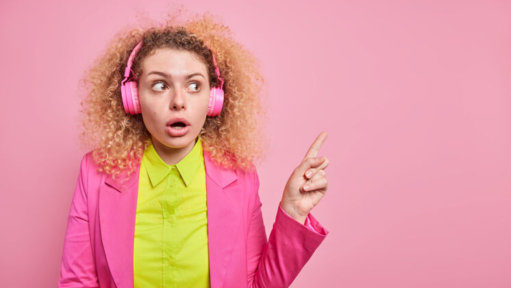 手指年轻的惊讶的卷发年轻的欧洲女人通过耳机听音乐保持嘴巴张开从右上角穿着正式的衣服隔离在粉红色的墙上女孩快乐人