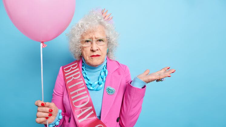 祖母摄影棚拍摄的满脸皱纹的白发老太太抬起手掌神情严肃 身着喜庆服装手持充气气球庆祝生日戒指丝带蛋糕