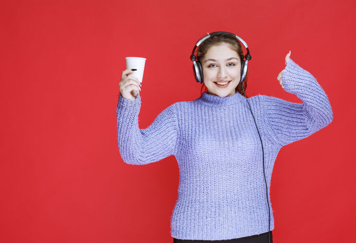 产品戴着耳机的女孩拿着咖啡杯 展示着享受的标志年轻人播放列表娱乐