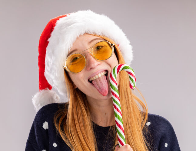 微笑微笑的年轻漂亮女孩戴着圣诞帽 戴着眼镜 抱着和舔着隔离在白墙上的圣诞糖果女孩舔年轻