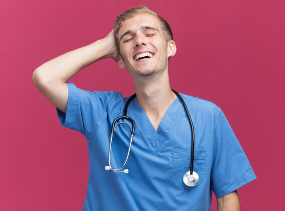 男性微笑着闭上眼睛年轻的男医生穿着医生制服听诊器把手放在头上隔离在粉红色的墙上人医生手