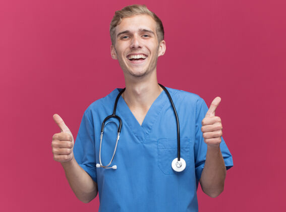 表情微笑着的年轻男医生穿着医生制服 听诊器在粉红色的墙上孤立地竖起大拇指制服医生站着