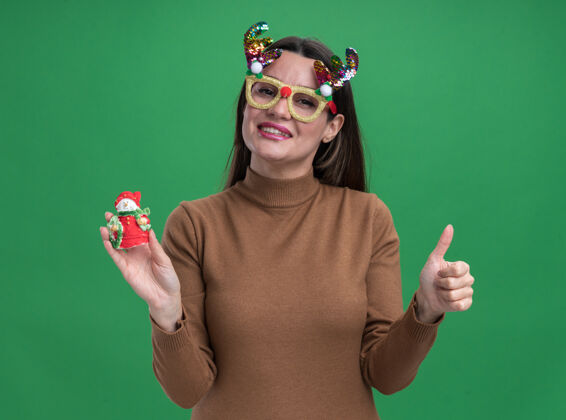 微笑微笑着的年轻漂亮女孩 穿着棕色毛衣 戴着圣诞眼镜 手里拿着圣诞玩具 在绿色的墙上孤立地竖起大拇指年轻秀眼镜