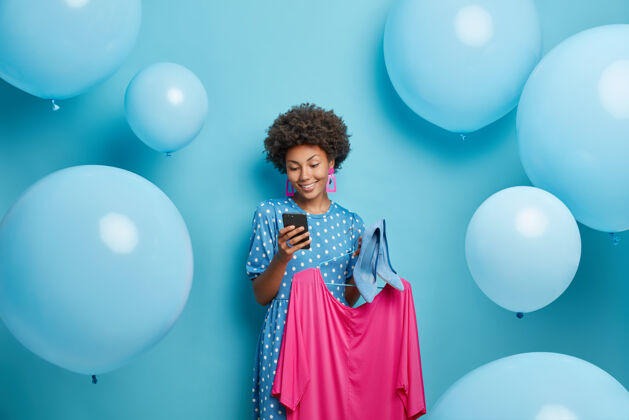手机女人通过智能手机发送短信 衣架上挂着粉色的裙子 高跟鞋在蓝色的衣架上为特殊场合做准备选择消费者美国