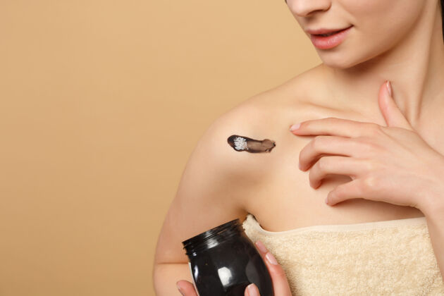 提升特写黑发半裸完美皮肤的女人 裸妆黑色面膜隔离在米色粉彩墙上手健康容器