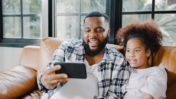 家庭快乐的非洲裔美国家庭爸爸和女儿在家里的沙发上玩得开心 用手机视频通话微笑爱科技