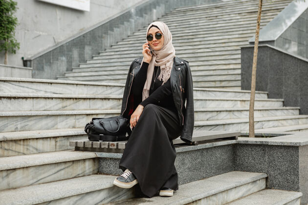 传统现代时尚的妇女戴着头巾 穿着皮夹克和黑色长袍 走在城市街道上用智能手机聊天阿拉伯语科技民族