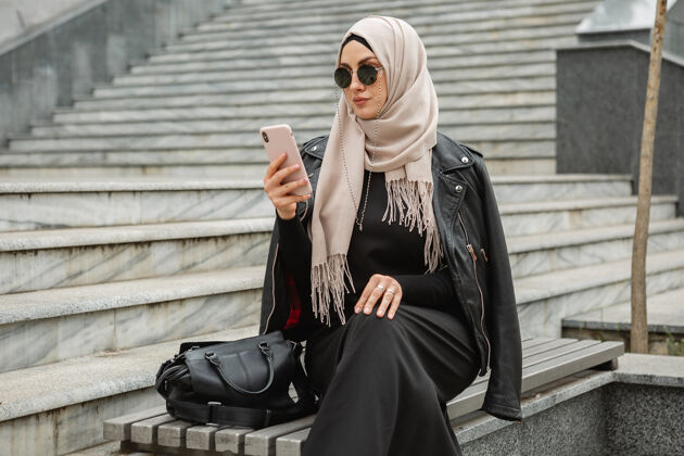 服饰现代时尚的妇女戴着头巾 穿着皮夹克和黑色阿巴亚在城市街道上用智能手机散步社交头巾街道