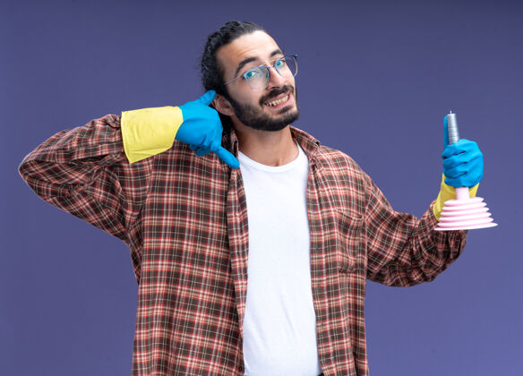 人微笑的年轻帅气的清洁工穿着t恤 戴着手套 拿着柱塞 在蓝色的墙上显示着电话的手势表演微笑市民