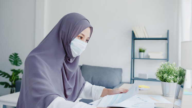 亚洲美丽的亚洲女士戴着口罩在客厅使用笔记本电脑和商业报告技术头巾