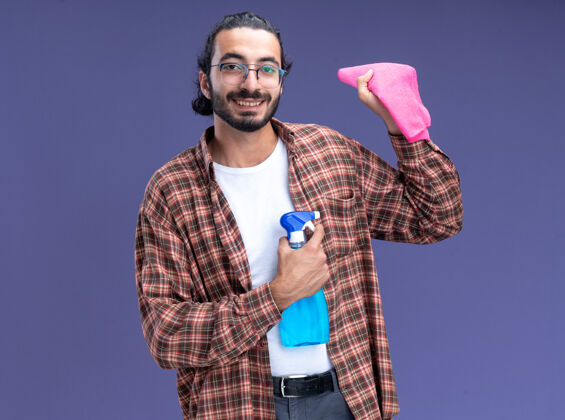 瓶子微笑的年轻帅气的清洁工穿着t恤拿着喷雾瓶用抹布隔离在蓝色的墙上破布年轻脸