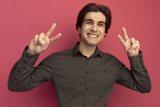 微笑面带微笑的年轻帅哥穿着黑色t恤 在粉红色的墙上显示出和平的姿态站着穿着人